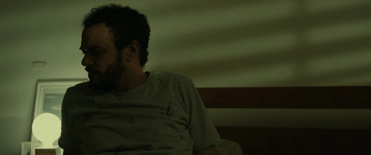 Terrified (2017) movie still: medium — A dimly lit room features a man sitting on a wooden headboard bed frame. Light from a nearby spherical table…