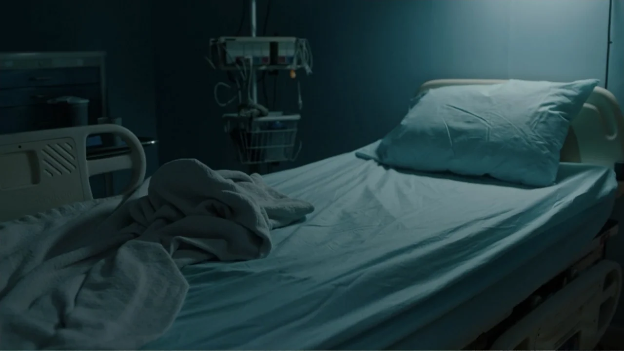 Sightless (2017) movie still: medium — A dimly lit hospital room contains an unmade medical bed with blue linens. A medical equipment stand is…