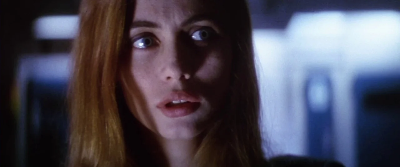 Mission: Impossible (1996) movie still: closeup — A dimly lit indoor corridor is visible with blue-toned fluorescent light sources. A young woman…