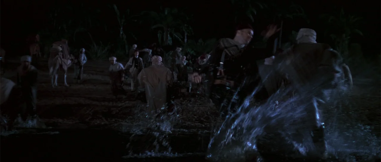 The Mummy (1999) movie still: fullbody — A group of people dressed in 1930s attire cross a river at night. One man in the foreground splashes…