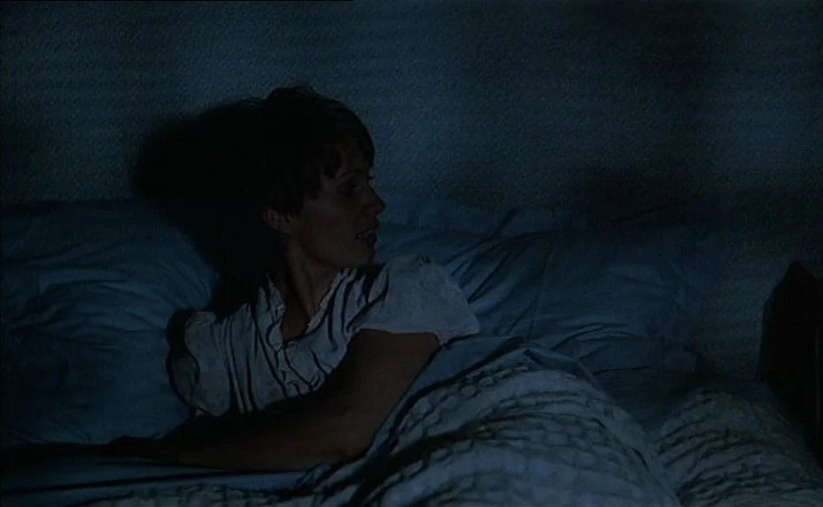 The Woman Next Door (1981) movie still: medium — A dimly lit bedroom features a person resting under soft blue-toned bedding. The subject lies on…