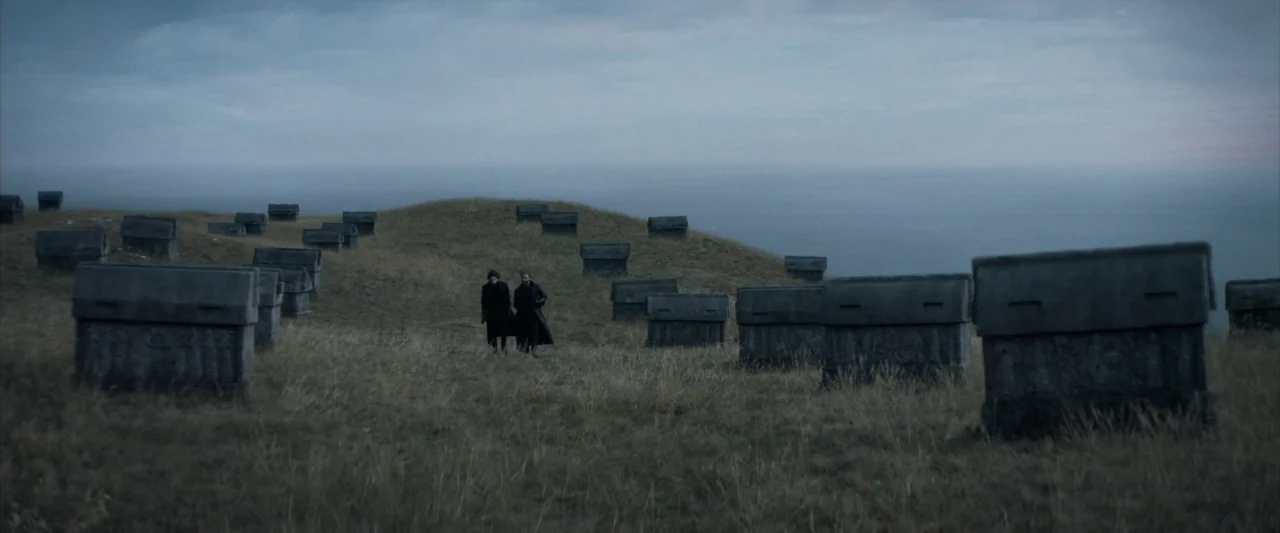 Dune (2021) movie still: wide — A vast grassy hill is covered with many evenly spaced stone box-like monuments. Two men in dark coats walk across…