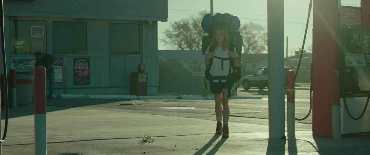 Wild (2014) movie still: fullbody — A paved gas station lot at midday with light glare and sparse infrastructure. A young woman walks toward the…