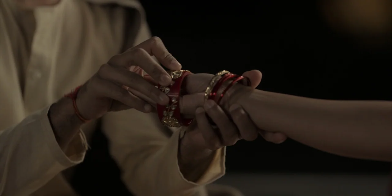 Dry Day (2023) movie still: closeup — A close-up view of two pairs of hands in a darkened room. One person places a decorative red bangle onto the…