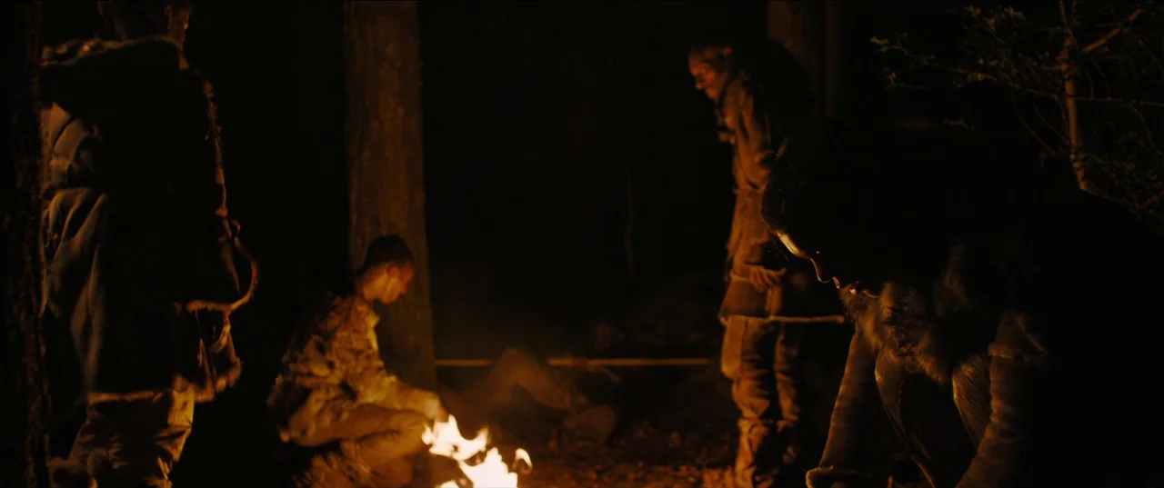 Out of Darkness (2022) movie still: medium — Four figures sit in a dark, nocturnal woodland area around a small, burning campfire. The individuals…