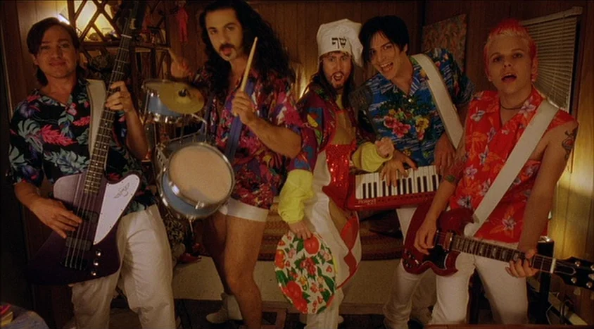 Hedwig and the Angry Inch (2001) movie still: medium — Five men stand in a room with wood-paneled walls and a venetian blind. The men wear…