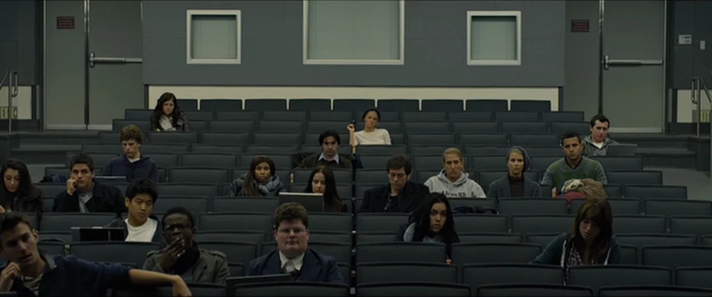 The Social Network movie still: wide — A spacious, modern university lecture hall with tiered grey rows of connected desk seats. A diverse group of…