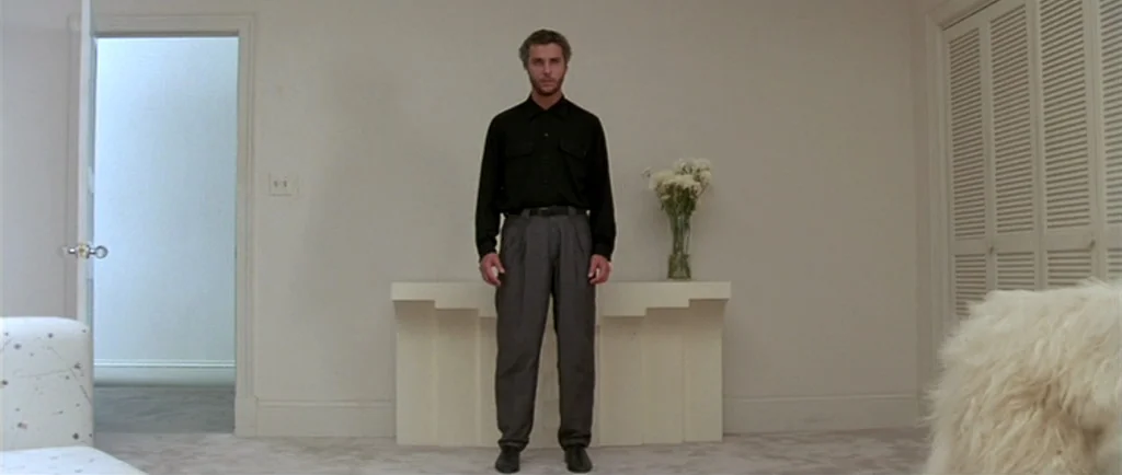 Manhunter (1986) movie still: fullbody — A man stands in the center of a white-walled room with minimalist white furniture. He remains perfectly…