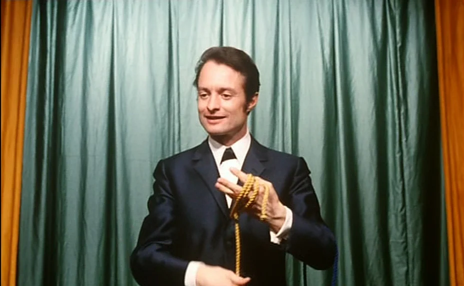 Stolen Kisses (1968) movie still: medium — A man stands in front of a draped teal curtain framed by gold side curtains. He performs a magic trick by…