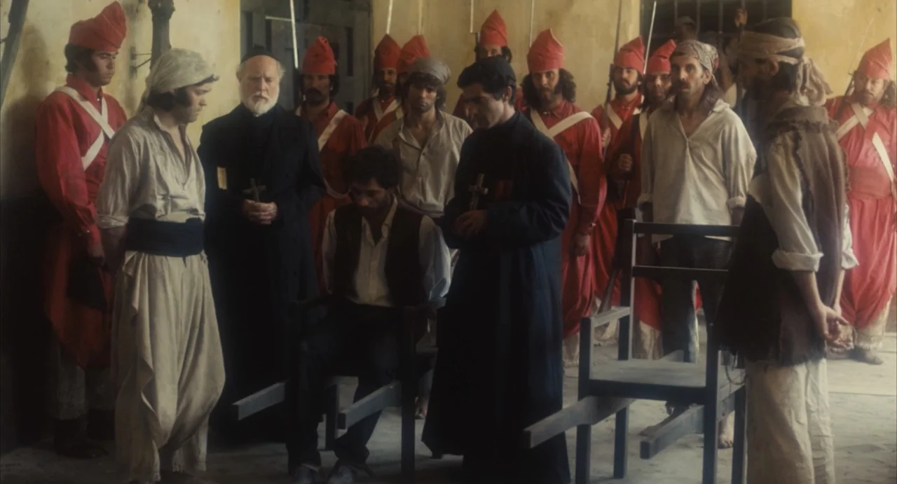 Camila (1984) movie still: medium — A stone-walled interior courtyard contains a group of soldiers and clergy members. A man sits in a wooden chair…