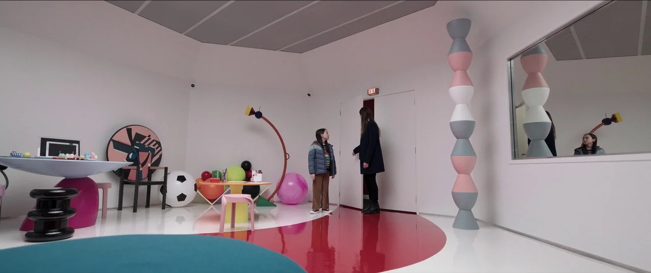 M3gan (2022) movie still: wide — A stark white minimalist room features rounded walls and bright red floor accents. A child and an adult woman stand…