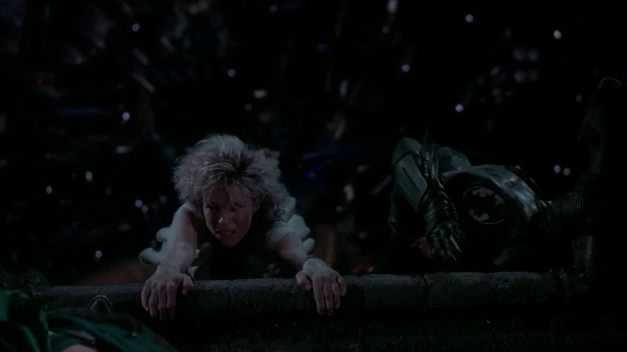 Batman (1989) movie still: medium — A dark rooftop edge at night overlooks a distant cityscape with glowing pinpoints of light. A blonde woman and a…
