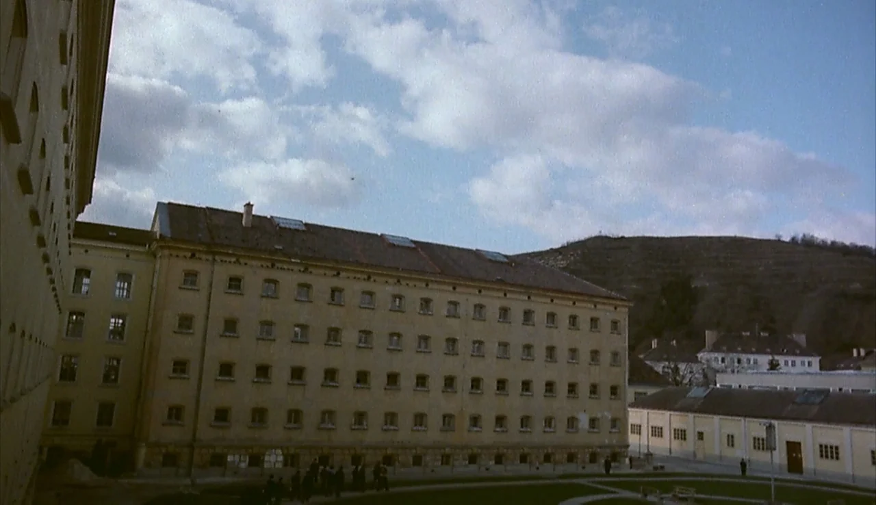 Angst (1983) movie still: establishing — A large multi-story stone prison building occupies the center of a courtyard. A rocky hill rises behind the…