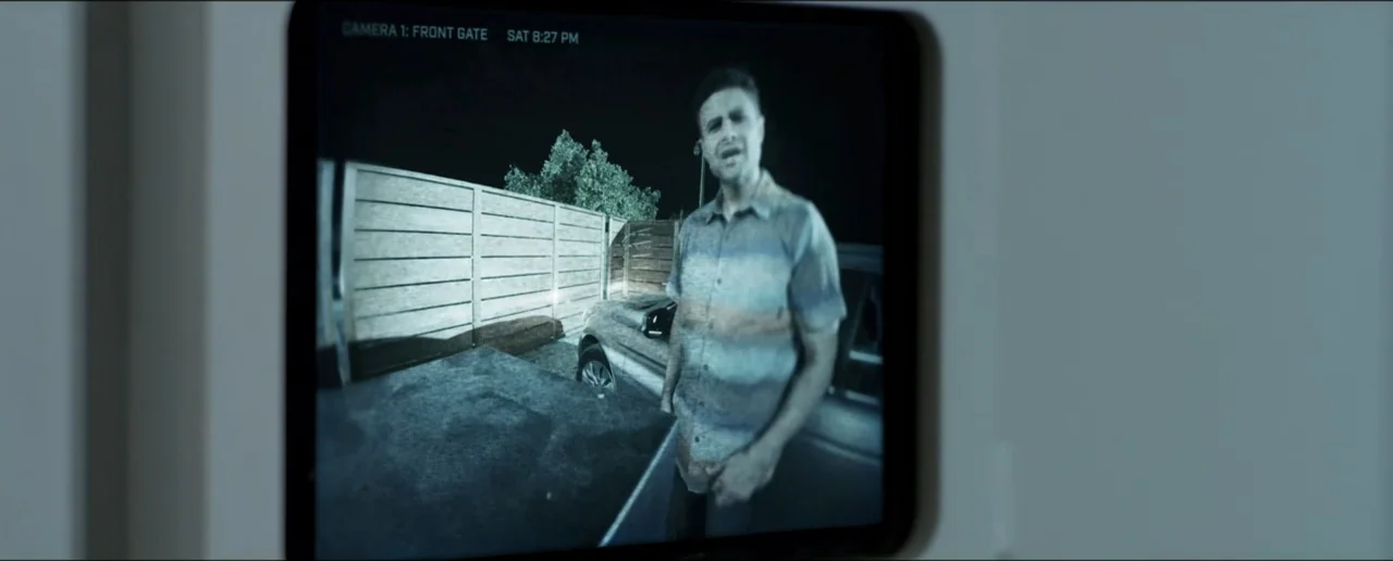 Hostages (2017) movie still: medium — A dark outdoor driveway illuminated by artificial light shows a wooden fence and car. A man in a striped shirt…