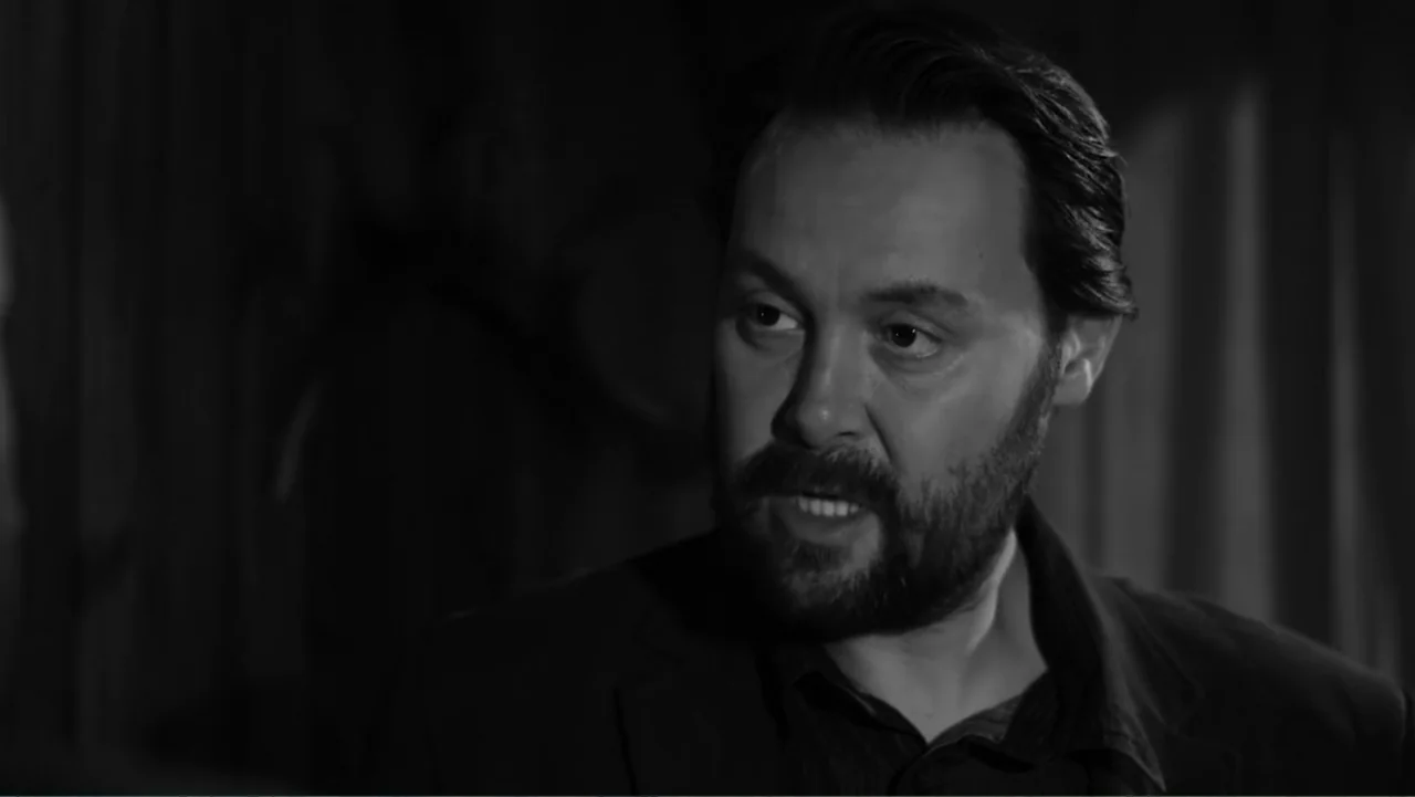 Creditors (2025) movie still: closeup — A dimly lit indoor space with textured curtain backdrop and dark shadow-filled corners. A man with a dark…