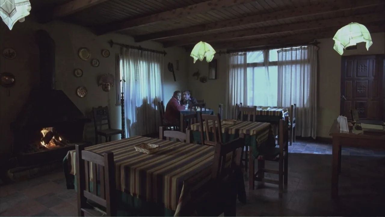 A Distant Place (2020) movie still: wide — A rustic dining room features wooden ceiling beams and several tables with striped tablecloths. Two…
