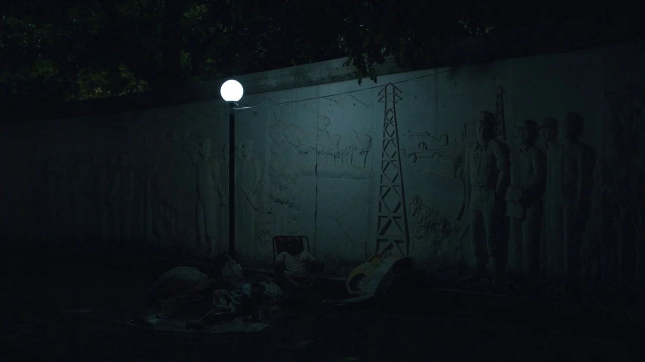 Cemetery of Splendour (2015) movie still: wide — A nighttime street corner features a concrete wall with bas-relief sculptures of workers. A single…
