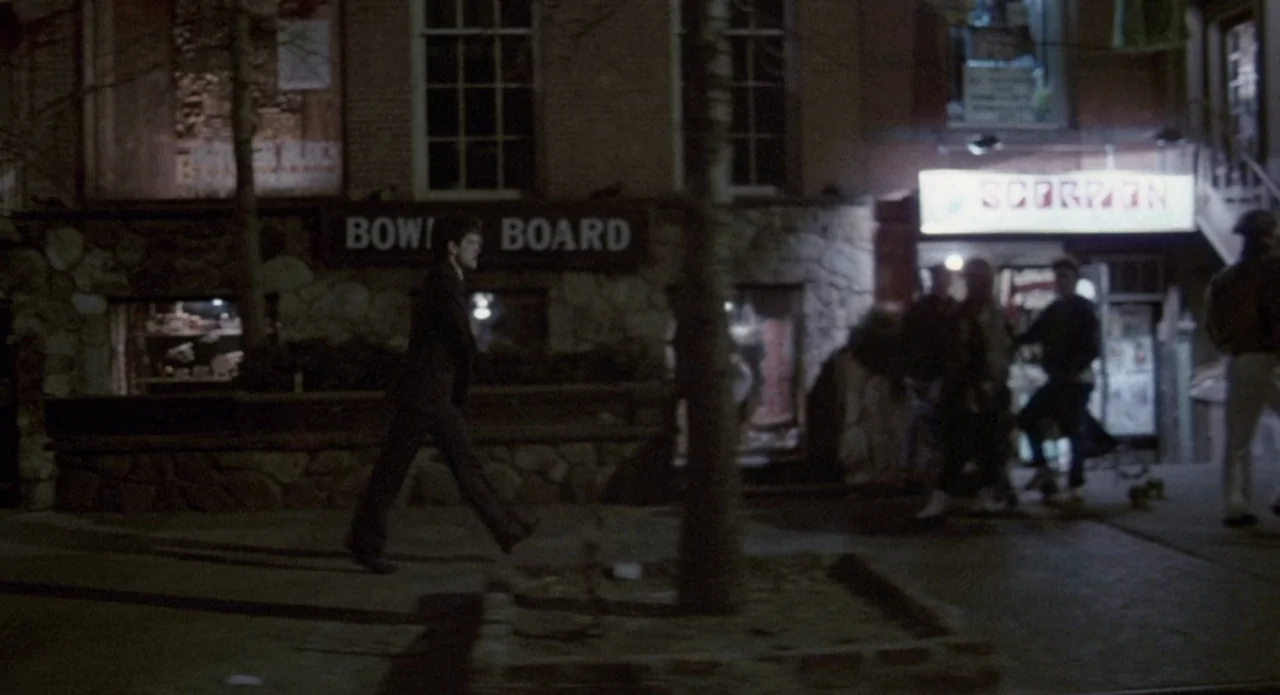 Brain Damage (1988) movie still: wide — A night view of a commercial building with stone accents and storefront windows. A man in a dark suit walks…