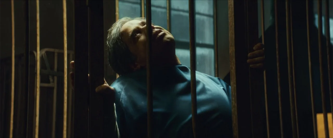 American Carnage (2022) movie still: medium — A dimly lit room contains a set of vertical metal bars obstructing the view. A man in a blue shirt…