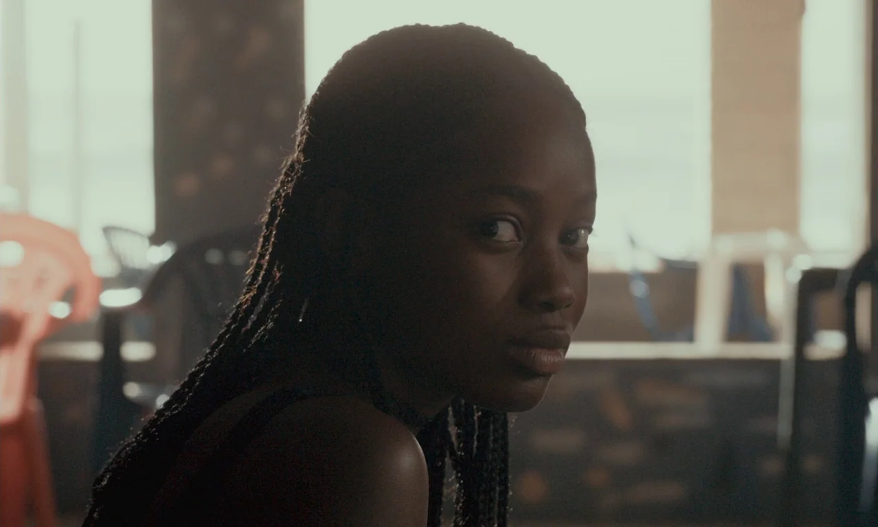 Atlantics (2019) movie still: closeup — A sunlit room features blurred chairs and tables in the background near a window. A young woman with long…