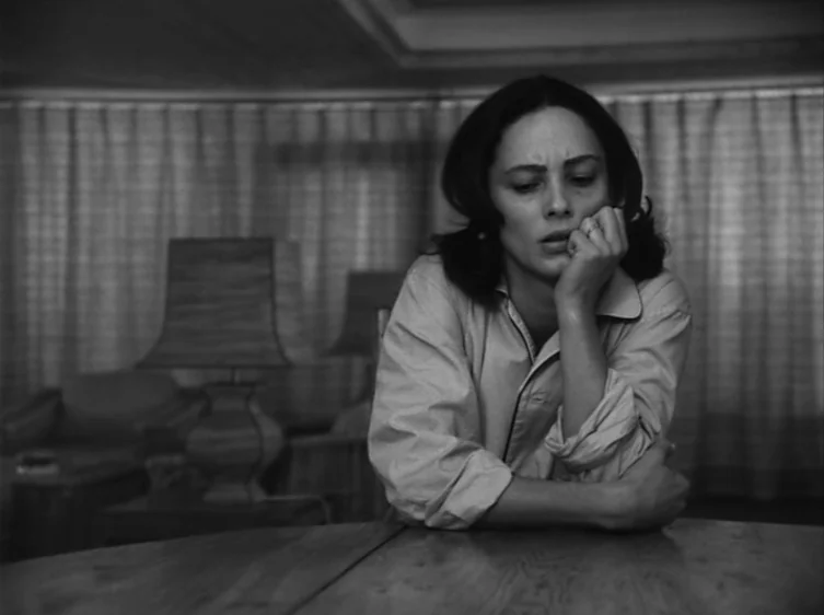 From the Life of the Marionettes (1980) movie still: medium — A dimly lit room with floor-to-ceiling curtains and a wooden table in front. A young…