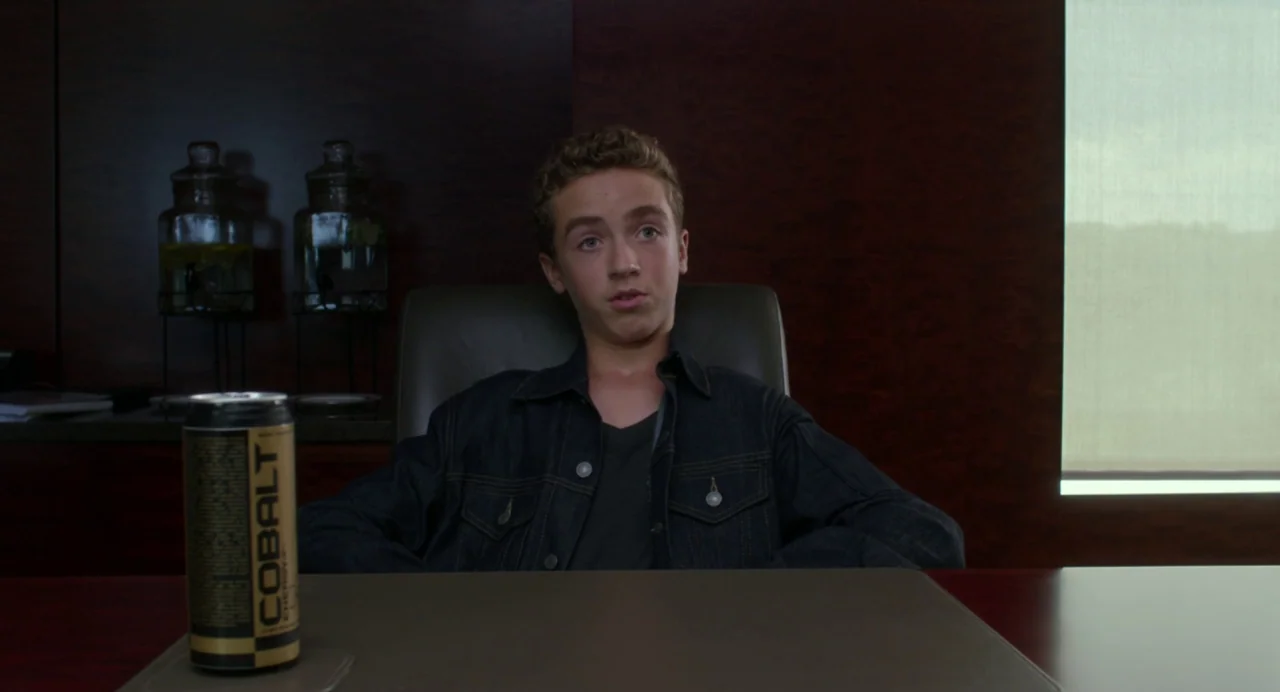 Maps to the Stars (2014) movie still: medium — A dimly lit room with dark wood paneling features a desk and office chair. A young man sits in the…