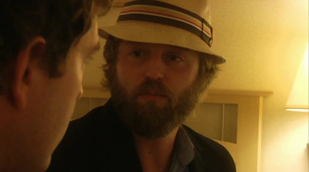 Humpday (2009) movie still: medium — An interior shot shows a man in a fedora talking to another person. The lighting comes from a warm lamp placed…