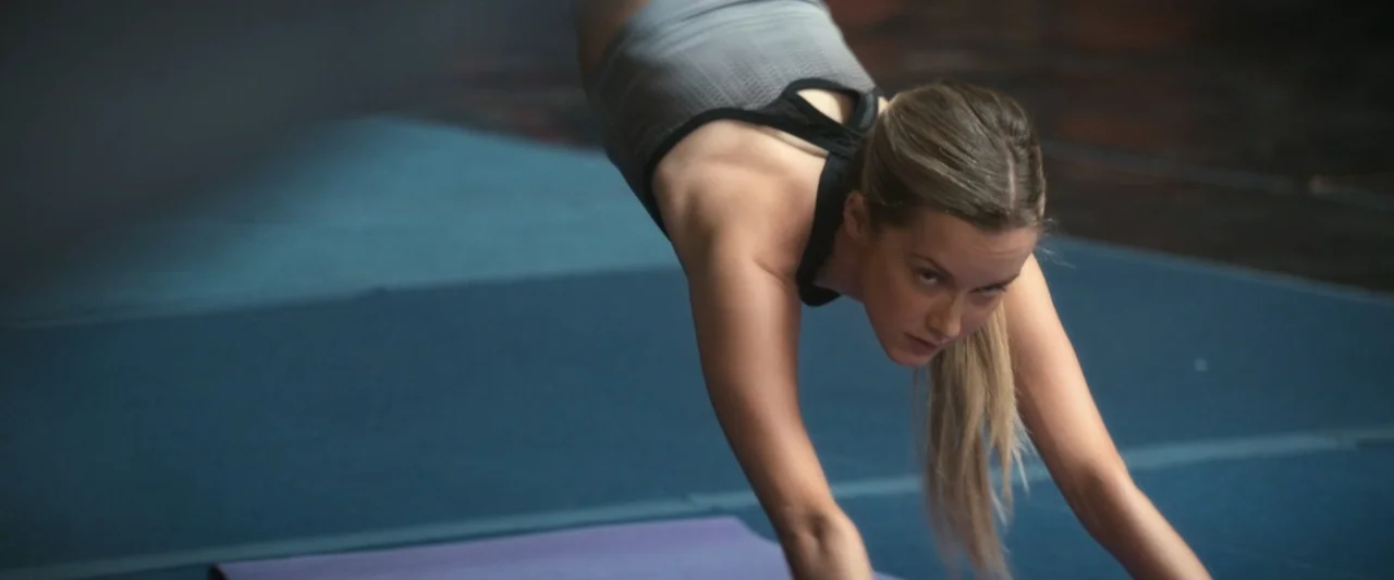 I Love America (2022) movie still: medium — A spacious athletic facility contains a blue padded floor and a purple yoga mat. A young woman with…