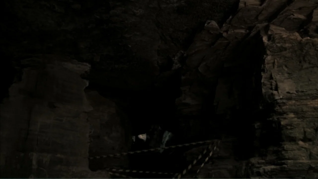 The Grotto (2022) movie still: establishing — A dark, rocky cave interior with textured walls and a small opening. A yellow and black hazard tape is…