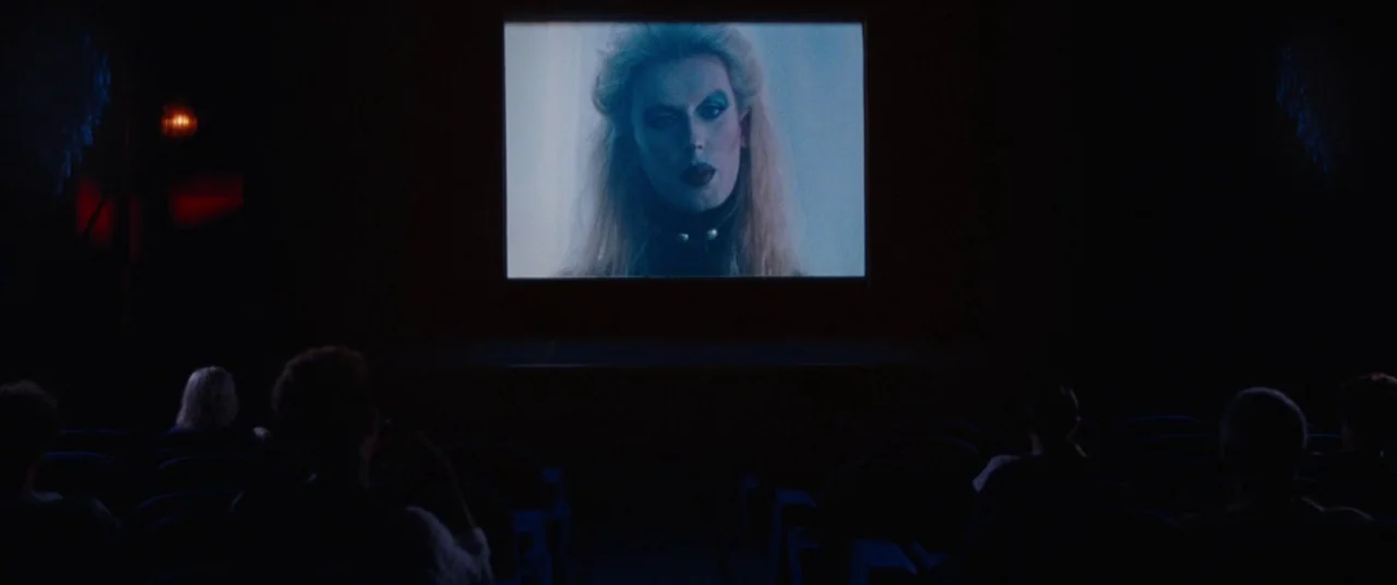 Knife+Heart (2018) movie still: wide — A dark movie theater with audience silhouettes watching a screen. A large close-up of a woman appears on the…