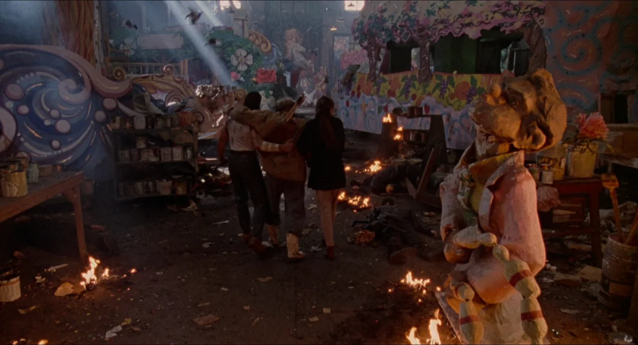 Hard Target (1993) movie still: fullbody — A cluttered workshop is filled with painted props and debris on the floor. Three people walk away…