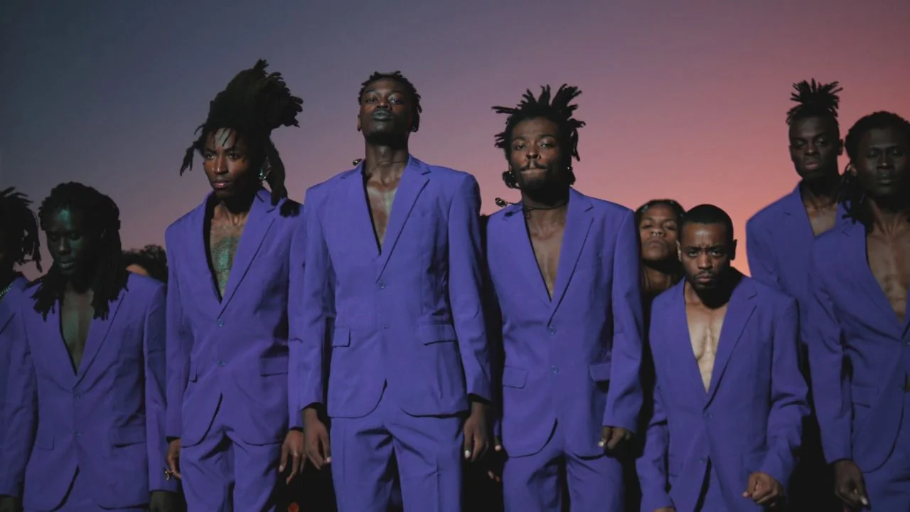 Black is King (2020) movie still: fullbody — A group of Black men in identical purple suits stand together in an open field. The background consists…