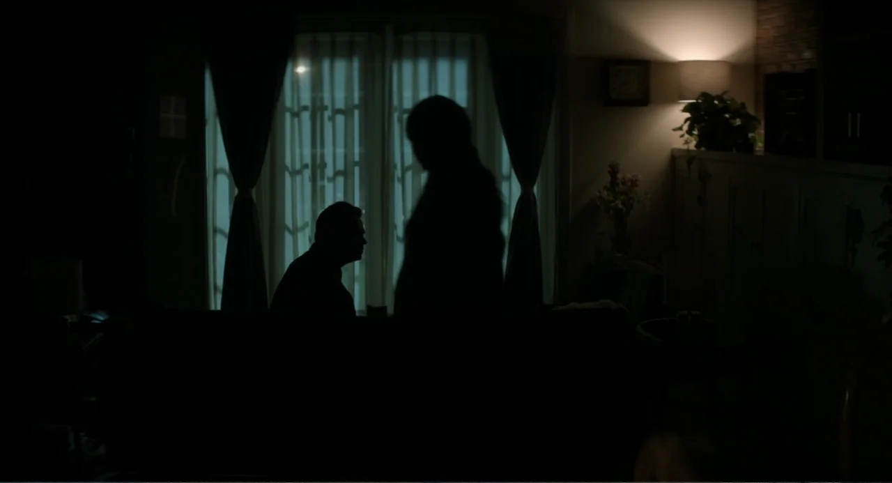 A Million Miles Away (2023) movie still: medium — A dimly lit room at night features a window covered by sheer curtains. Two individuals stand in…