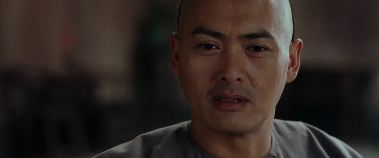 Crouching Tiger, Hidden Dragon (2000) movie still: closeup — A man stands in a dimly lit, sparsely furnished indoor space. The man looks forward,…