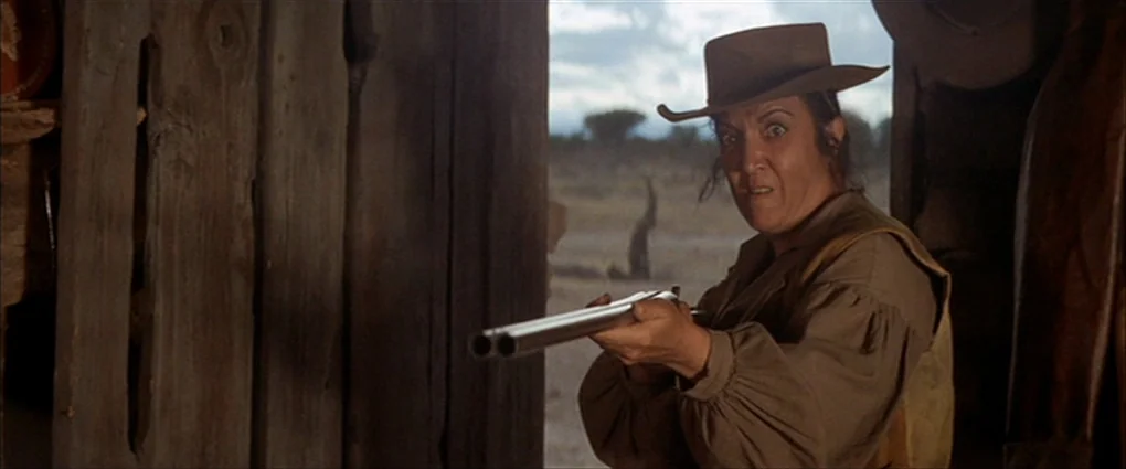 Pat Garrett & Billy The Kid (1973) movie still: medium — A rustic barn doorway looks out toward a dry, overcast landscape. A woman aims a…