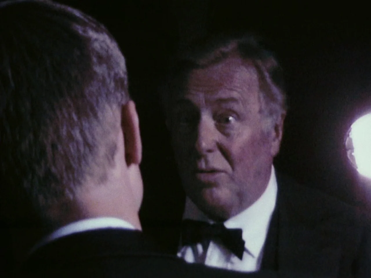 Festen (1998) movie still: closeup — A dimly lit room with two men dressed in formal evening attire. The older man stands facing the younger man…