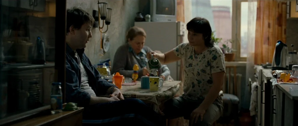 Elena (2011) movie still: medium — A cluttered apartment kitchen with a dining table and aging appliances. A woman pours tea from a teapot while a…