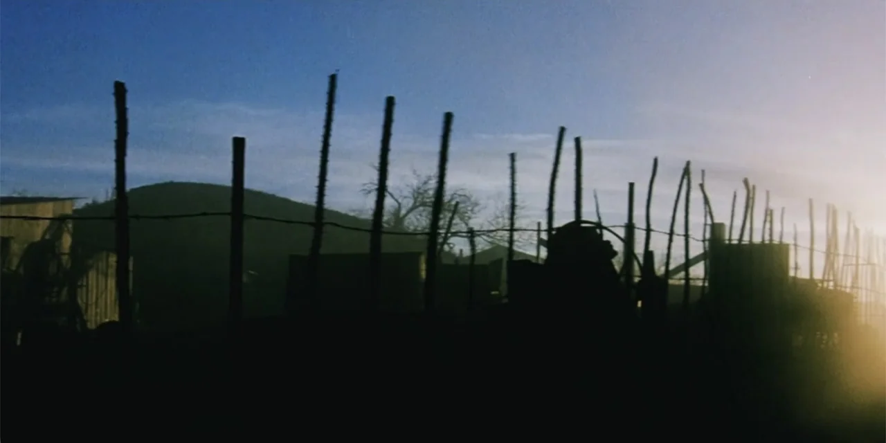 El sótano (2024) movie still: wide — A row of vertical wooden fence posts stands against a backdrop of distant hills. Sunlight flares from the right…