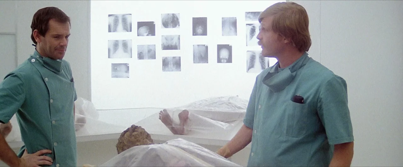 The Beyond (1981) movie still: medium — A stark white clinical room contains autopsy tables covered by translucent plastic sheets. Two men in green…