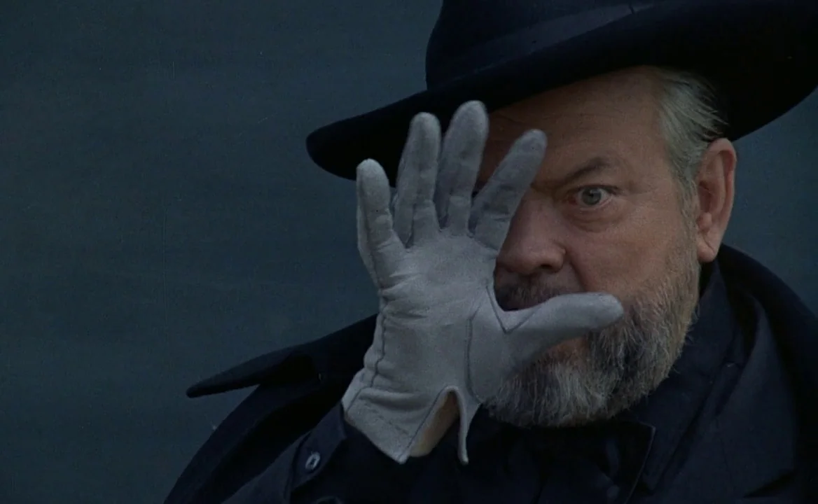 F for Fake (1973) movie still: closeup — An older man stands against a dark, featureless background wearing a black hat. He raises a grey gloved…