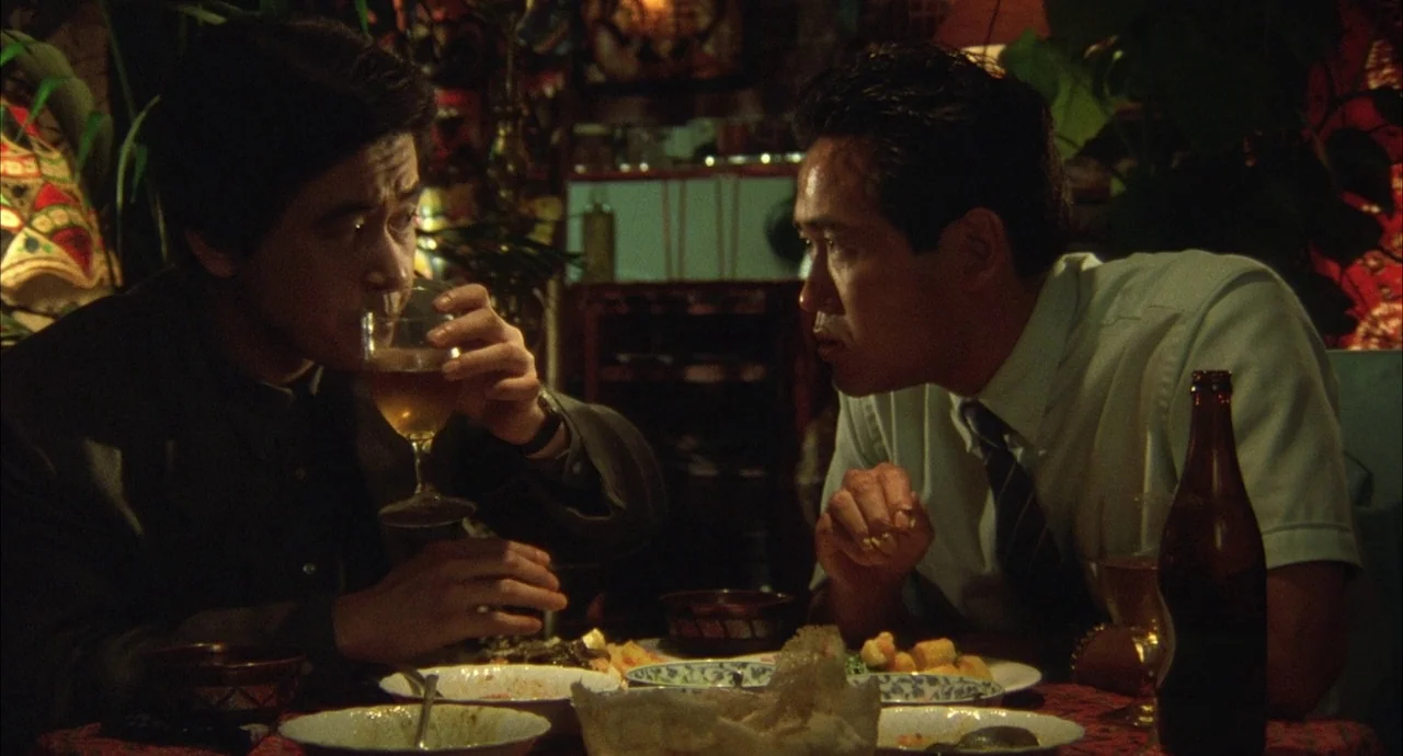 The Discarnates (1988) movie still: medium — Two men sit at a table covered in plates and bottles in a restaurant. They lean in toward each other…