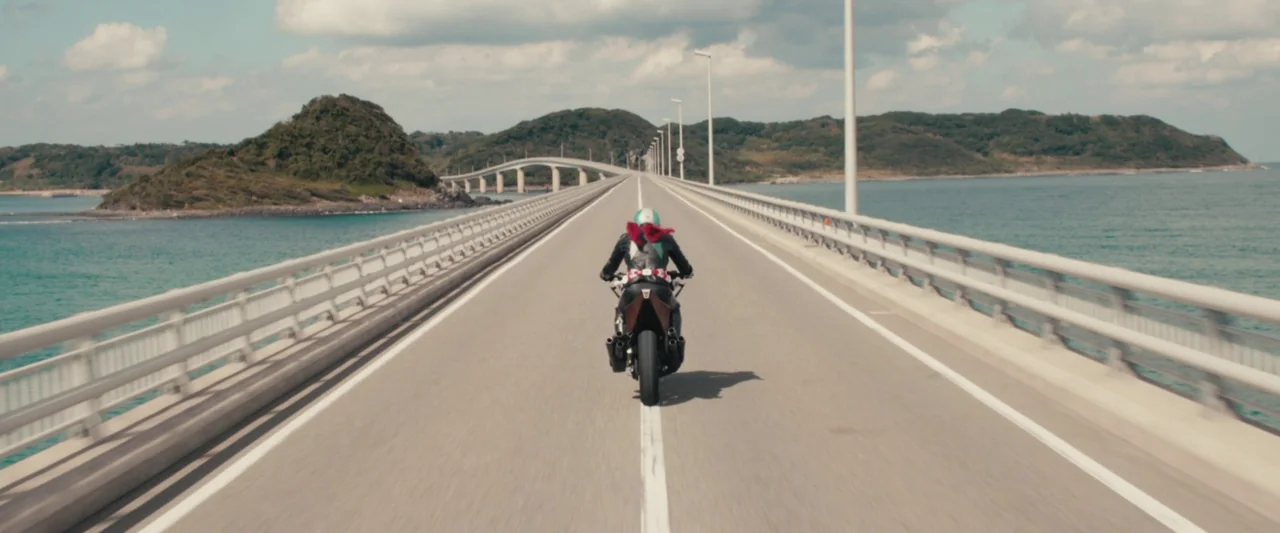 Shin Kamen Rider (2023) movie still: wide — A long paved bridge extends across open water between distant landmasses. A person on a motorcycle rides…