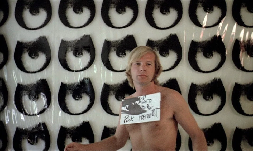 Lions Love (1969) movie still: medium — A white wall covered in repeating black eye graphics serves as a background. A shirtless man stands in front…