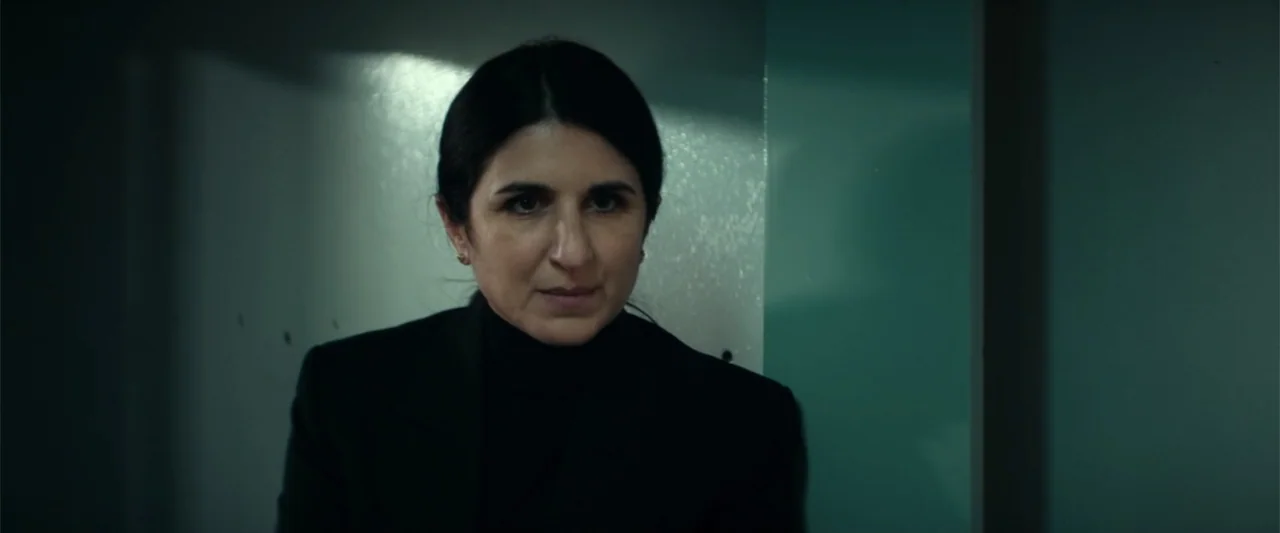 McWalter (2025) movie still: closeup — A dimly lit industrial hallway features smooth teal-colored wall panels and metallic surfaces. A woman in…