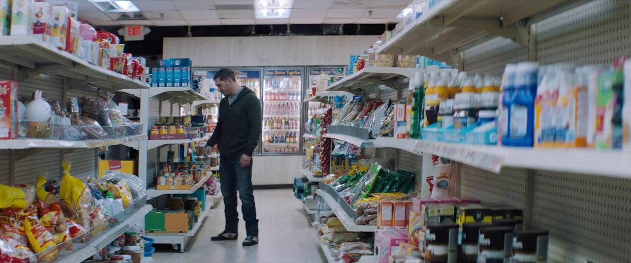 Venom (2018) movie still: wide — A quiet convenience store aisle filled with stocked shelves of various food products. A man in a dark hoodie stands…