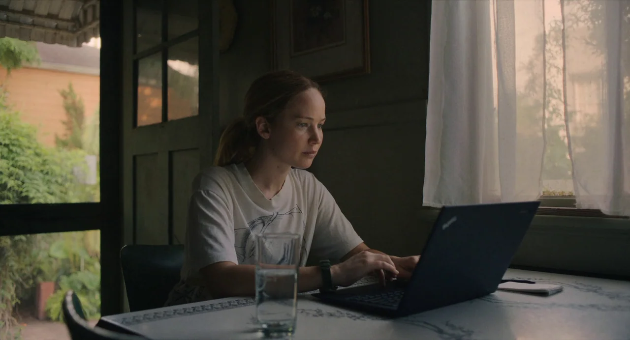 Causeway movie still: medium — A woman sits at a table near a window with thin white curtains. She types on a dark laptop with a glass of water…