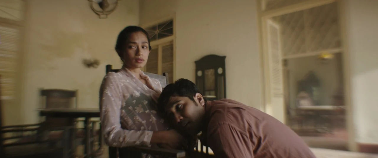 Impetigore (2019) movie still: medium — A spacious veranda with large doorways and vintage wooden furniture in a historic home. A man rests his head…