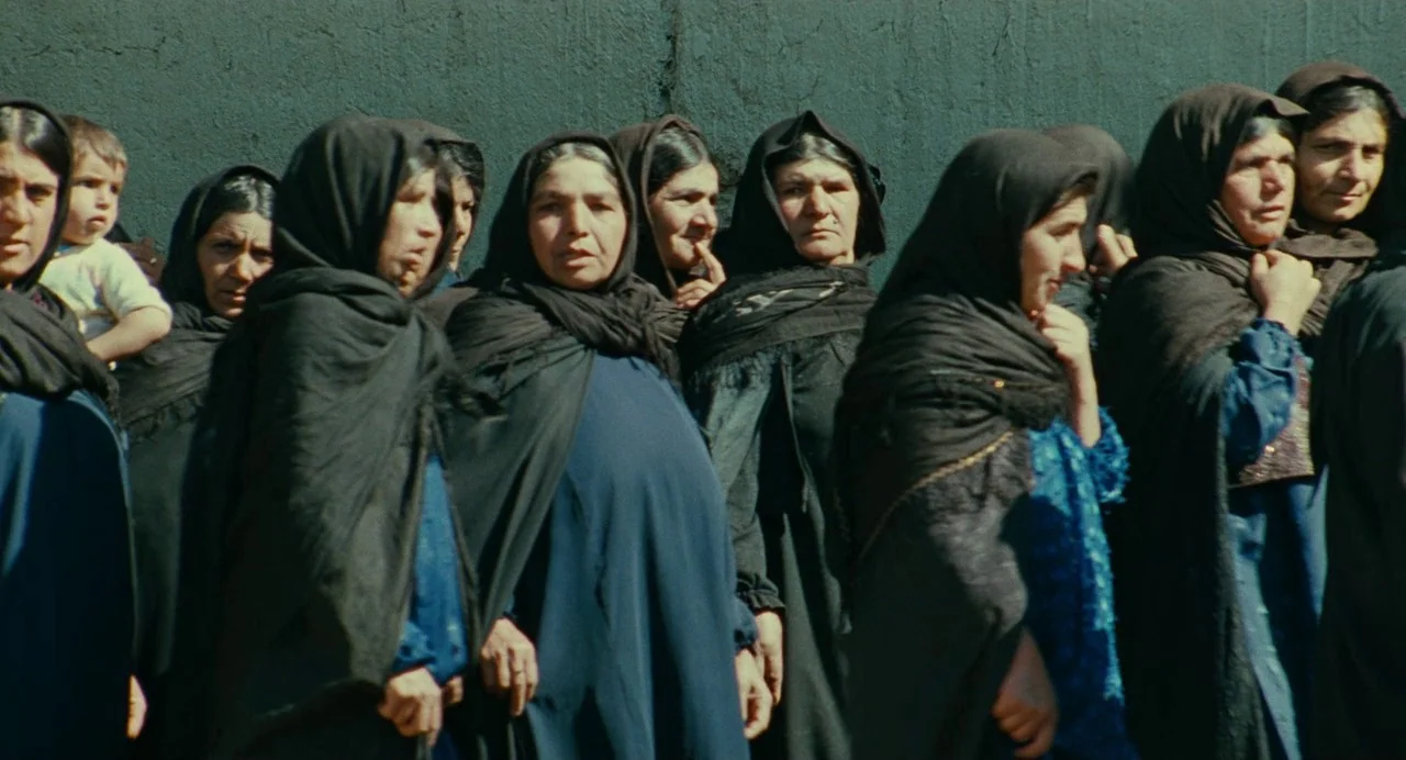 The Wind Will Carry Us (1999) movie still: medium — A group of women in dark headscarves stand in front of a textured concrete wall. They are…