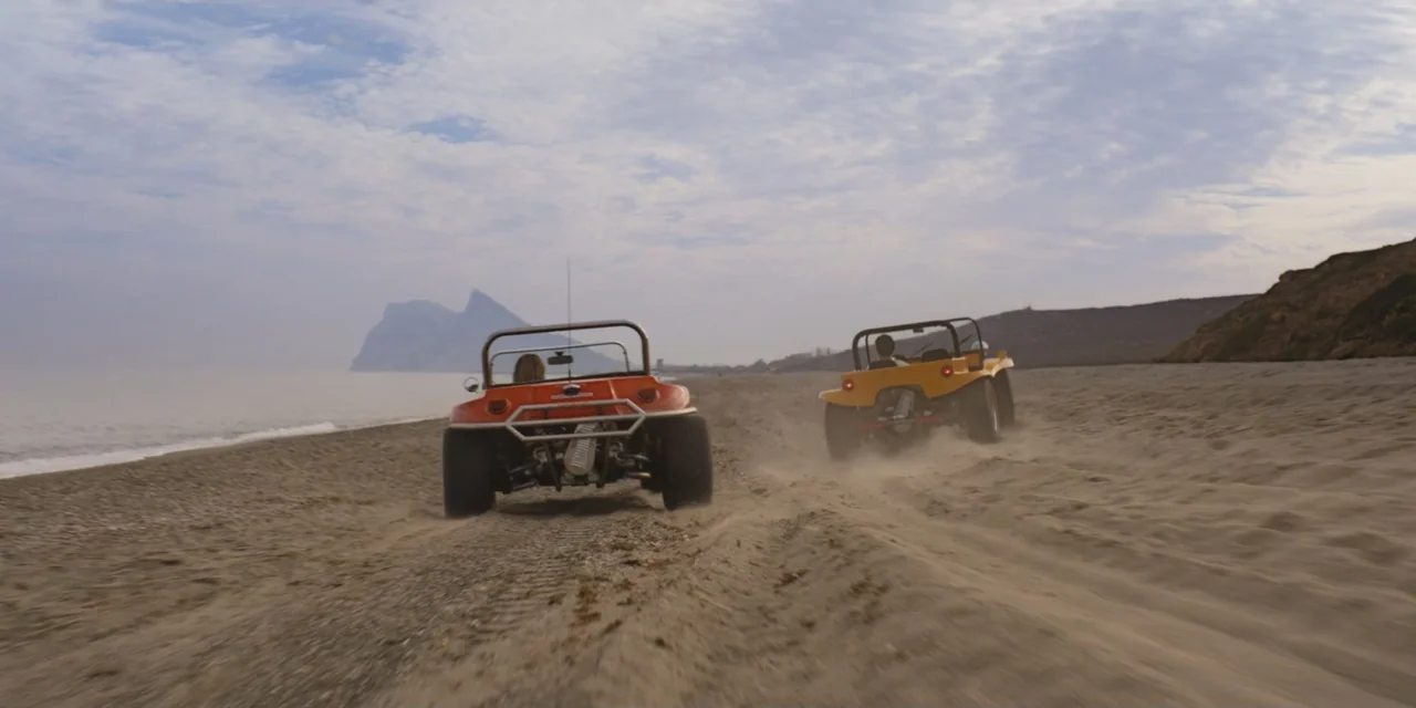 Your Fault (2024) movie still: wide — A wide beach with light sand stretches towards a distant coastal rock formation. Two colorful dune buggies…