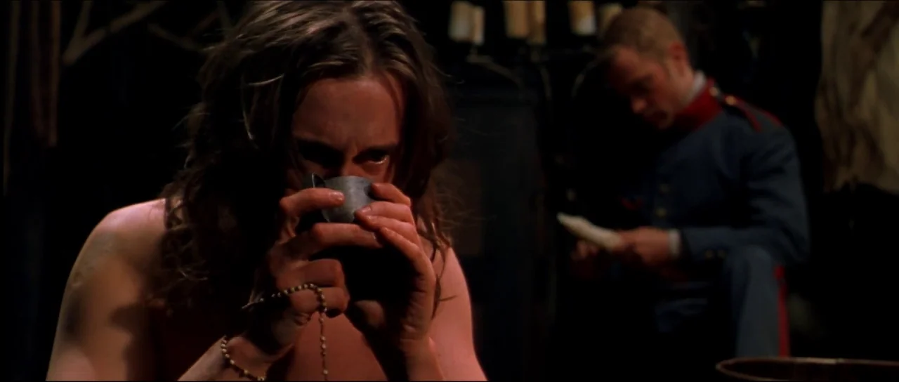 Ravenous (1999) movie still: medium — A dark, dimly lit barracks room features shadows and minimal furniture in the background. A shirtless man…